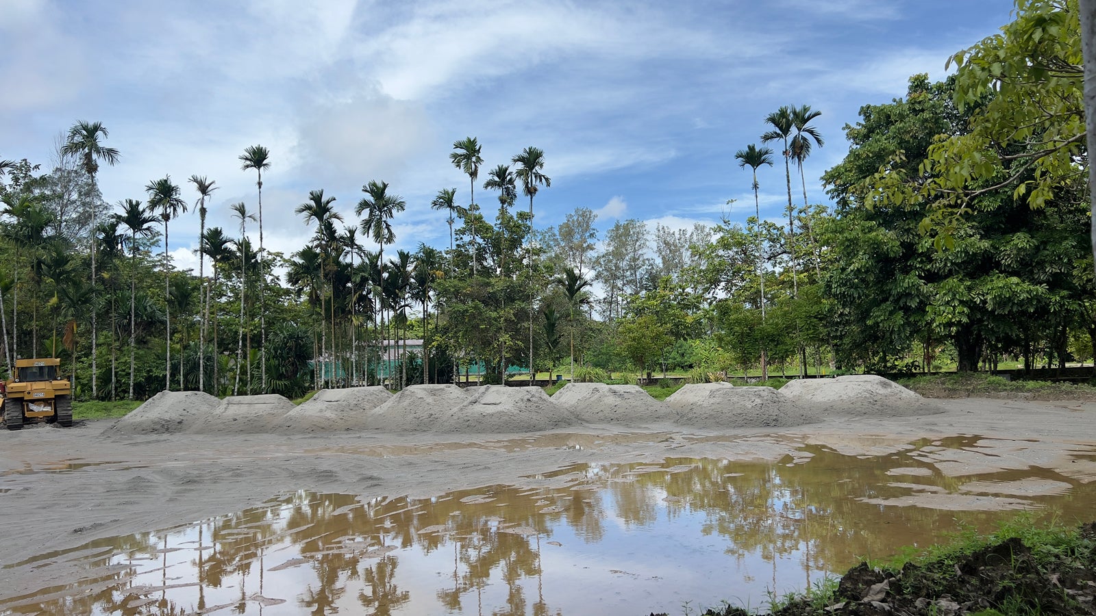A construction site with piles of sand under a cloudy sky. Reflective puddles are in the foreground. Tall trees, including several palm trees, are in the background. A yellow construction vehicle is partially visible on the left.