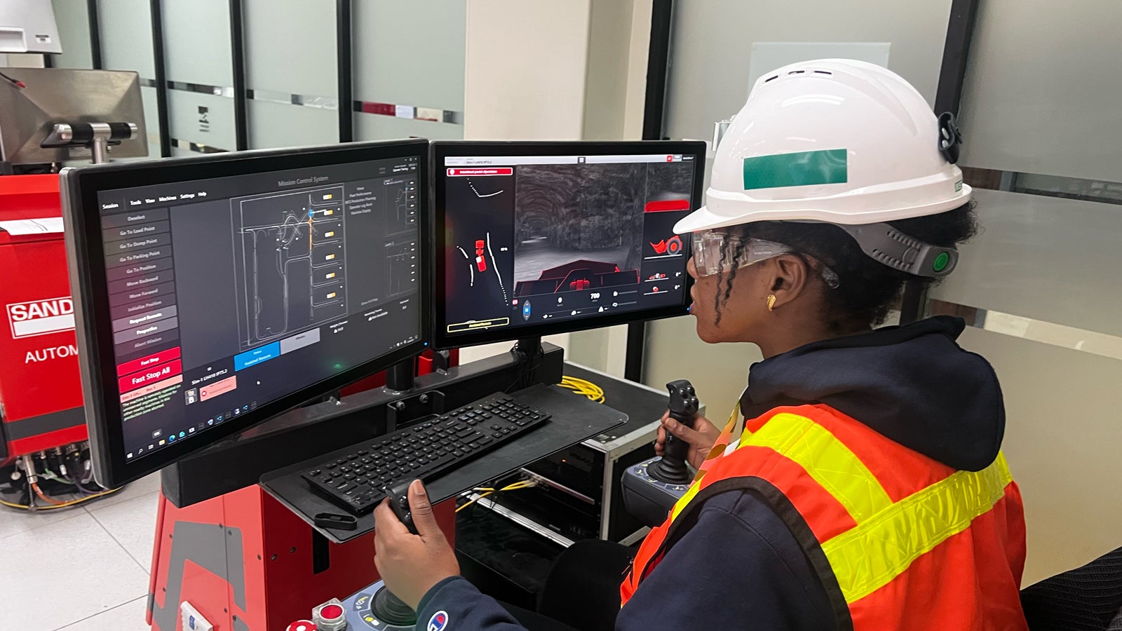 A person wearing protective gear, including a helmet and safety vest, operates a remote control system. Two monitors display technical data and visuals related to machinery or equipment control.