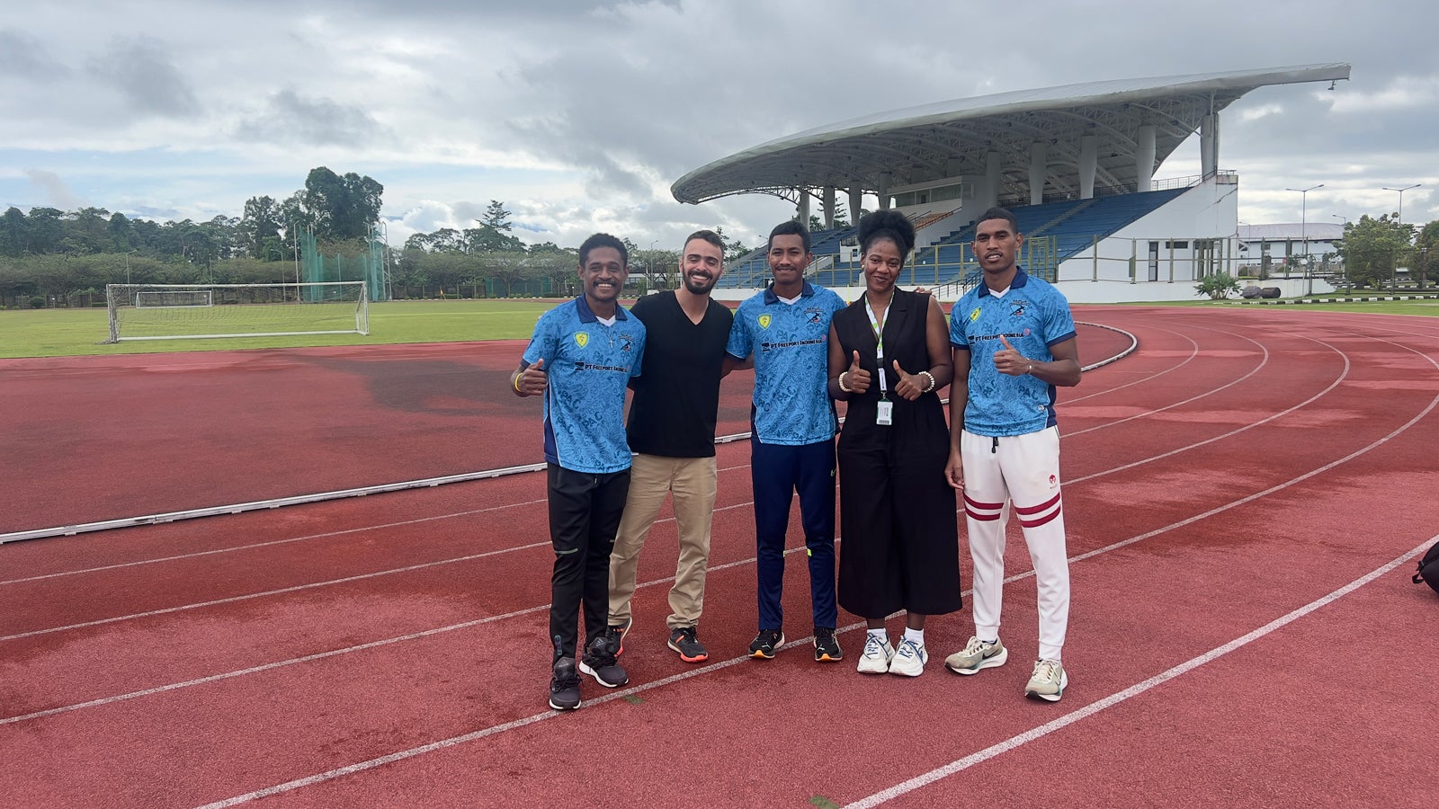 A group of five people stands on a red running track in an outdoor stadium. They are smiling and giving thumbs up. The background shows a large covered seating area and greenery under a cloudy sky. They appear to be dressed in athletic attire.