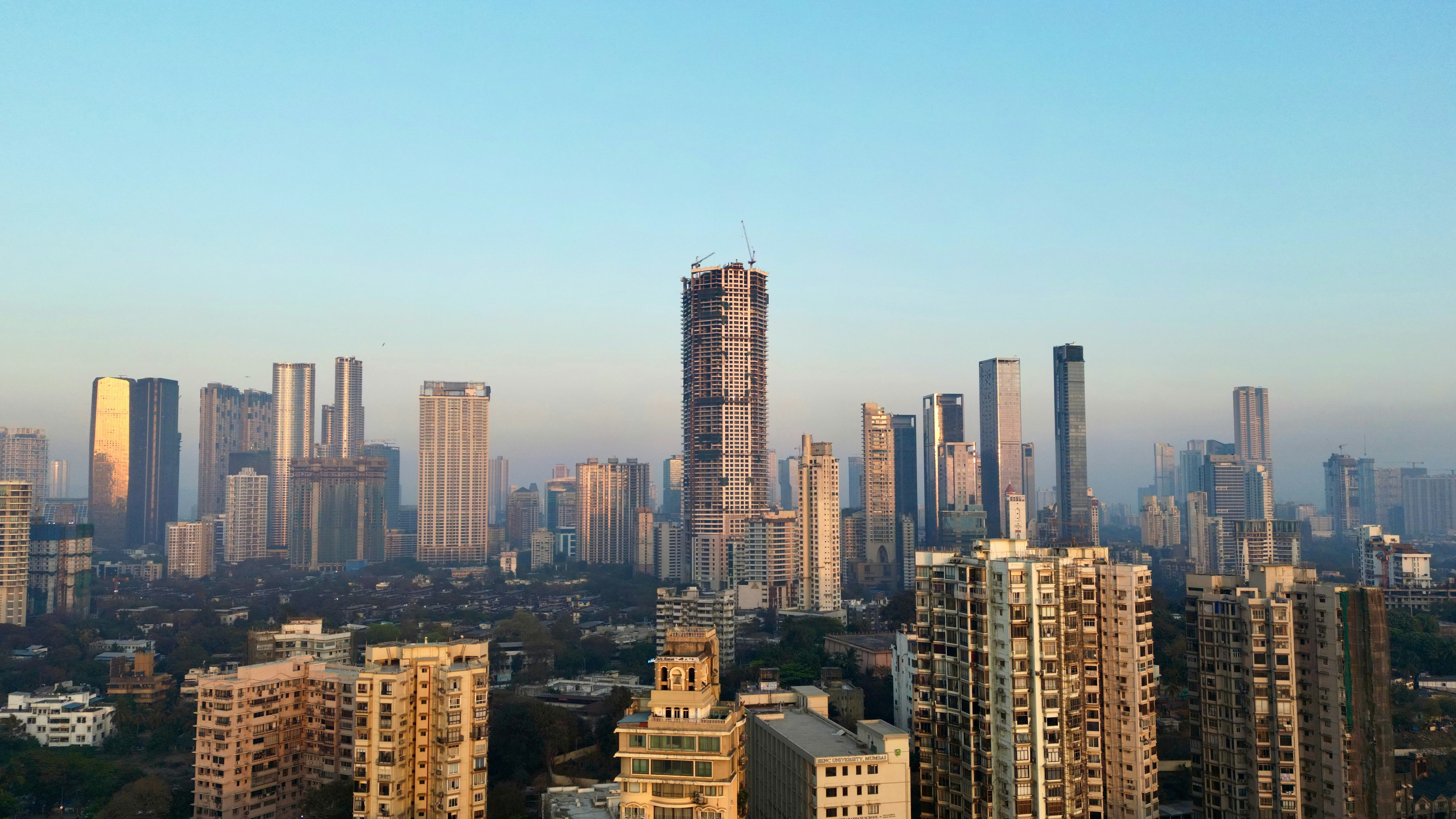 A panoramic view of a cityscape featuring numerous tall buildings under a clear blue sky. The skyline is dominated by modern skyscrapers, some under construction.