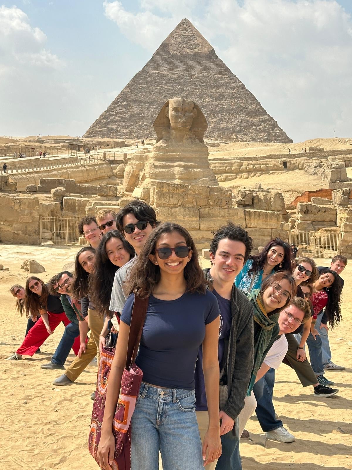 A group of people stands in front of the Sphinx and Great Pyramid of Giza. The individuals are smiling, and they are arranged in a line that curves slightly. The sky is partly cloudy, casting soft shadows over the ancient structures.