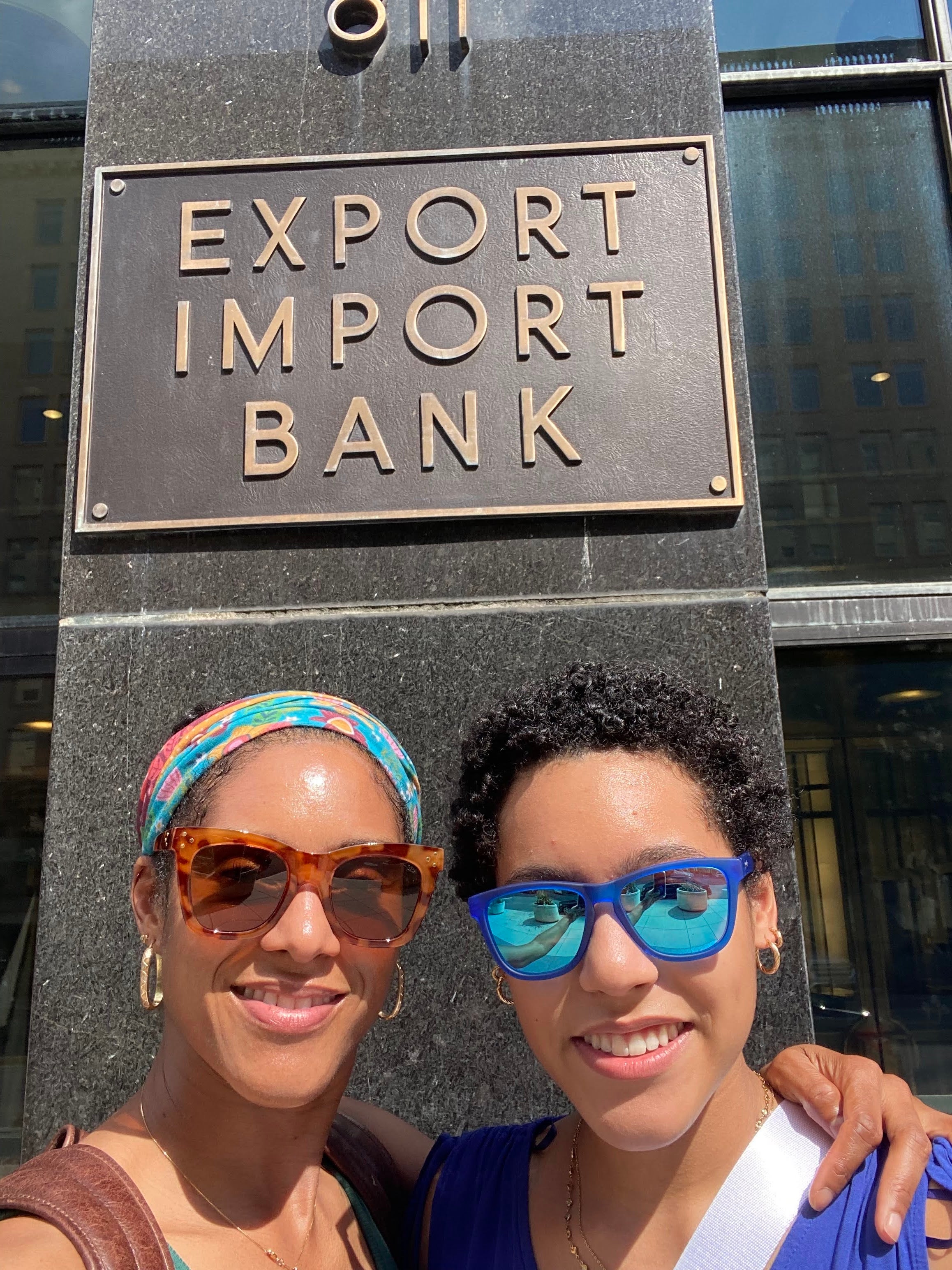 Two young women take a selfie in front of a sign for the Export Import Bank.