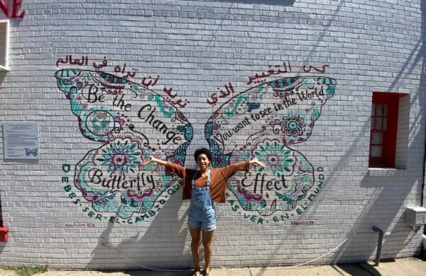 A young woman with short black hear spreads out her arms in front of a mural of butterfly wings.