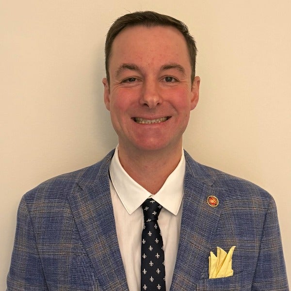 Alex Colas Des Francs, brown hair styled to side, light skin, slight smile, dark suit with striped shirt and tie