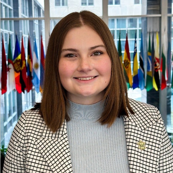 Ally Swindell, long brown hair, warm smile, gray turtleneck, checkered blazer, flags in background