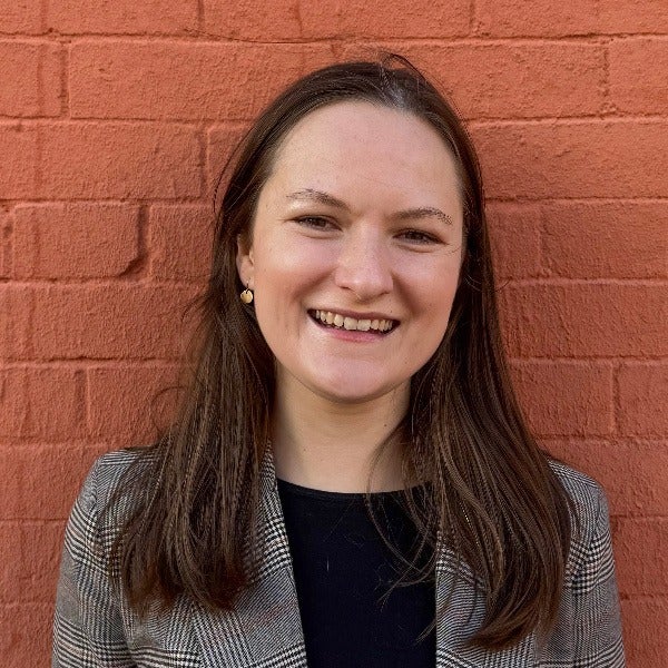 Alyssa Harvie, long brown hair, warm smile, small earrings, gray houndstooth blazer over black top, brick background