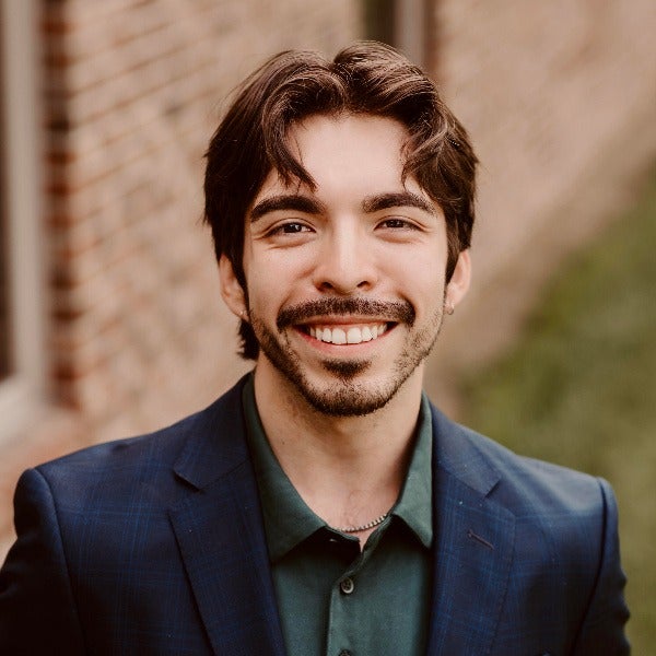 Andres Ayala, dark brown wavy hair, beard, warm smile, navy blazer over green collared shirt