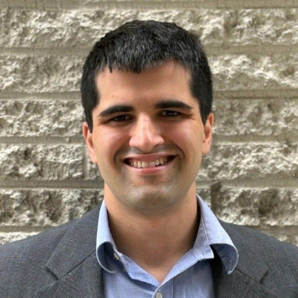 Antrim Lottick, short dark hair, warm smile, light blue shirt, gray blazer, textured brick background