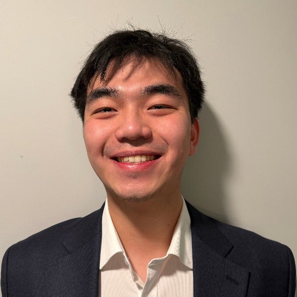 Auren Zhang, dark short hair, warm smile, navy blazer over white collared shirt, beige background