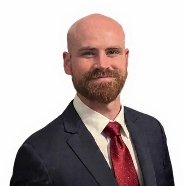 Austin Nicholson, bald, full brown beard, navy suit, red patterned tie, warm smile, white background
