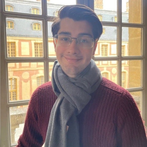 Carlos Aliseda, dark hair, glasses, gray scarf, burgundy sweater, slight smile, standing by window