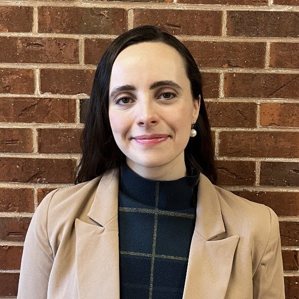 Cecilia Rike, long dark hair, fair skin, subtle smile, tan blazer over dark striped top, brick wall background