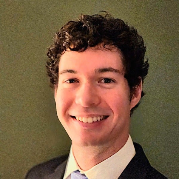 Daniel Budai, dark curly hair, bright smile, white shirt, dark suit jacket, olive background