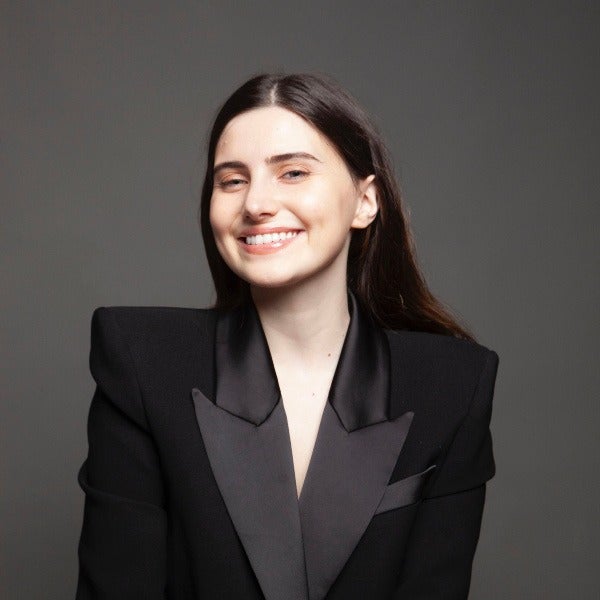 Elsa Alonso, long dark brown hair, bright smile, black blazer with satin lapels, gray background