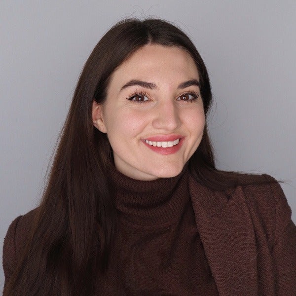 Emma Zubak, long dark brown hair, warm smile, brown turtleneck sweater, gray background