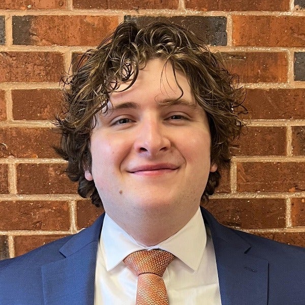 Gabriel Williams, brown curly hair, glasses, warm smile, blue suit, rust-colored tie, white shirt, brick background