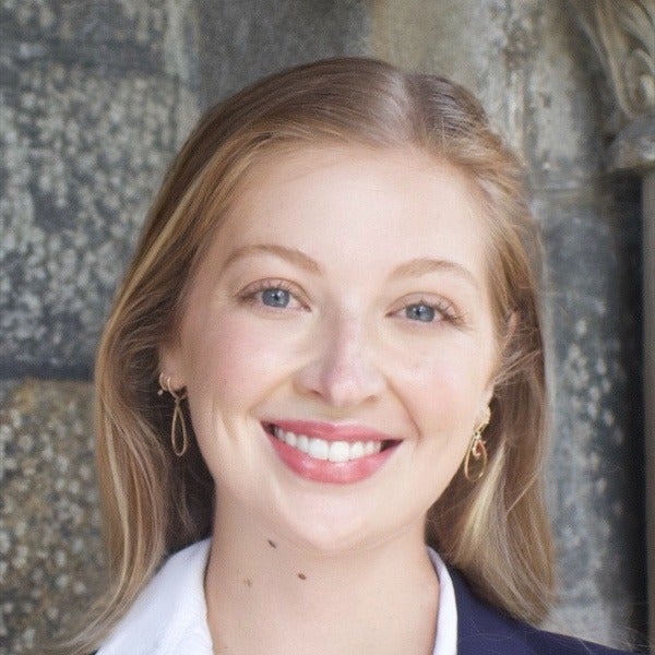 Georgia Gardner, shoulder-length blonde hair, blue eyes, warm smile, white collar under navy blazer, industrial background
