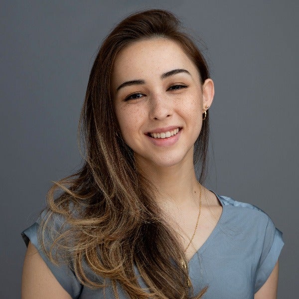 Gina Joerger, long brown hair with highlights, warm smile, light blue top, small hoop earrings, gray background