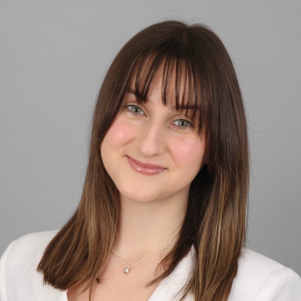 Grace Funk, brown hair with bangs, blue eyes, gentle smile, white top, pendant necklace, gray background