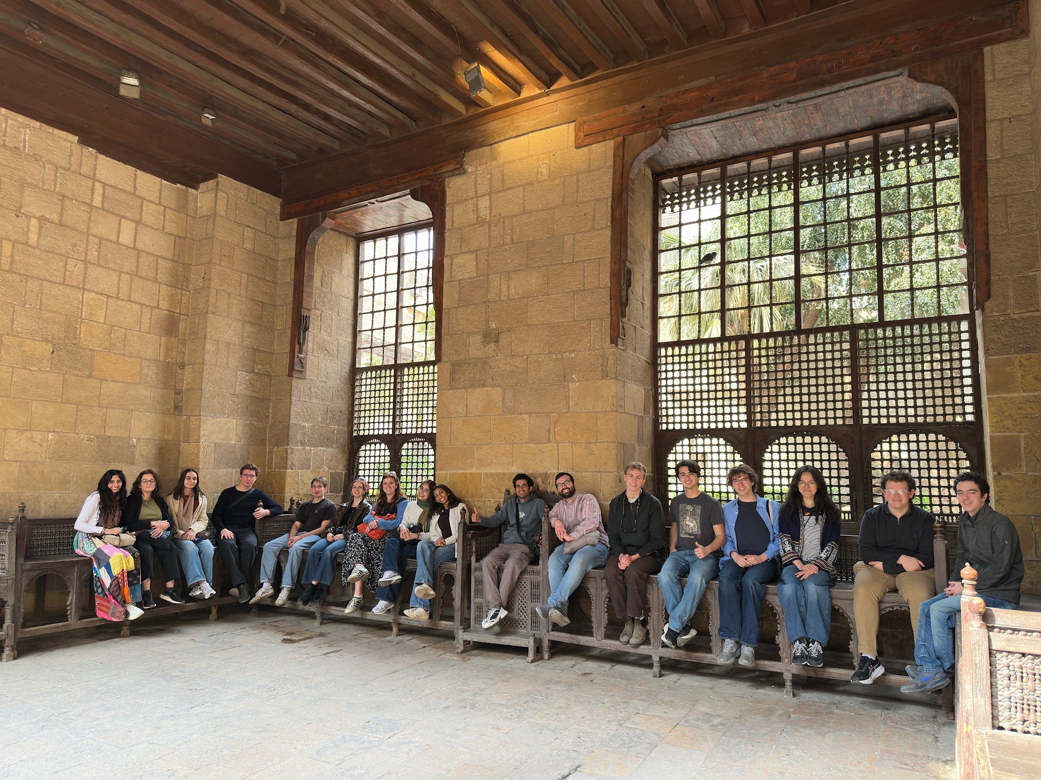 A large group of people sits on a long bench inside a historical stone building with high ceilings and wooden beams. Two large lattice windows are visible behind them, revealing a glimpse of greenery outside.