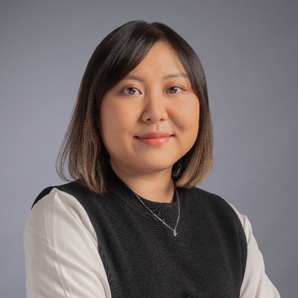 Hailey Chan, shoulder-length dark brown hair, gentle smile, black vest over white blouse, delicate necklace, gray background