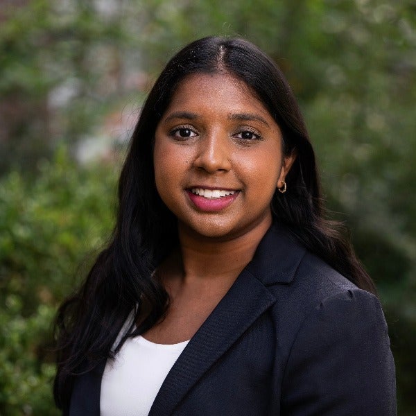 Hiran Dewar, long dark hair, warm smile, white top, black blazer, natural outdoor background