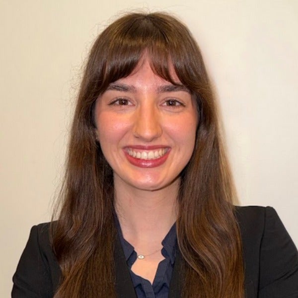 Isabella Bournas, long brown hair with bangs, warm smile, black blazer over dark blue top, beige background