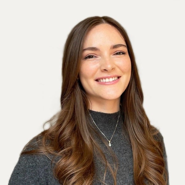 Jamie Schlegel, long brown wavy hair, bright smile, dark gray sweater, delicate necklace, light background