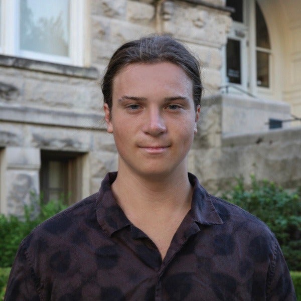 Jared Abazia, dark shoulder-length hair, light skin, slight smile, dark camouflage shirt, stone building background