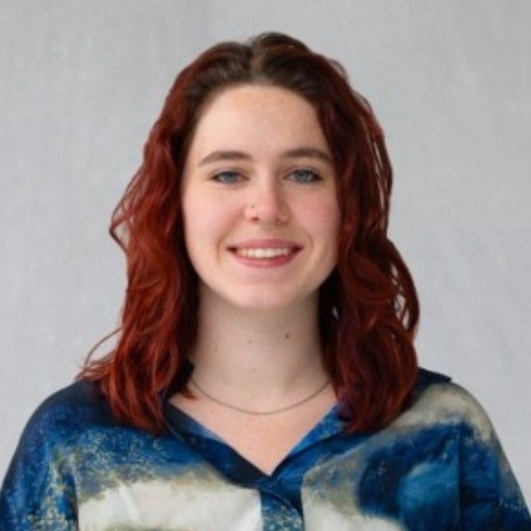 Katya Mouradian, shoulder-length reddish-brown wavy hair, blue eyes, warm smile, blue and white patterned shirt, light background