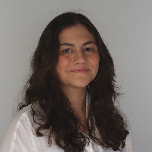 Laila Ghabrial, long dark wavy hair, warm smile, white collared shirt, light gray background