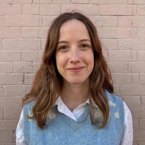 Lauren Eskra, wavy brown hair, warm smile, blue knit vest over white collared shirt, brick wall background