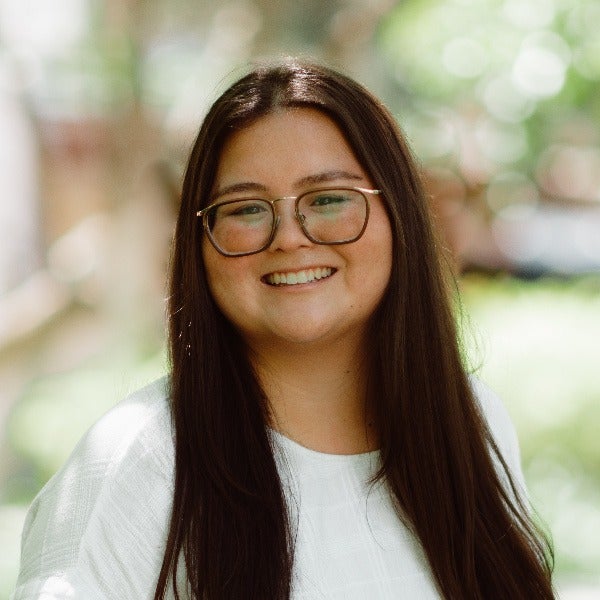 Lauren Yenari, long dark brown hair, glasses, bright smile, white shirt, blurred outdoor background