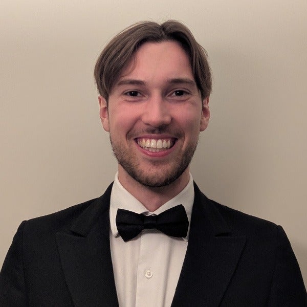 Lucien Obrien, brown center-parted hair, beard, warm smile, black tuxedo with bow tie, beige background