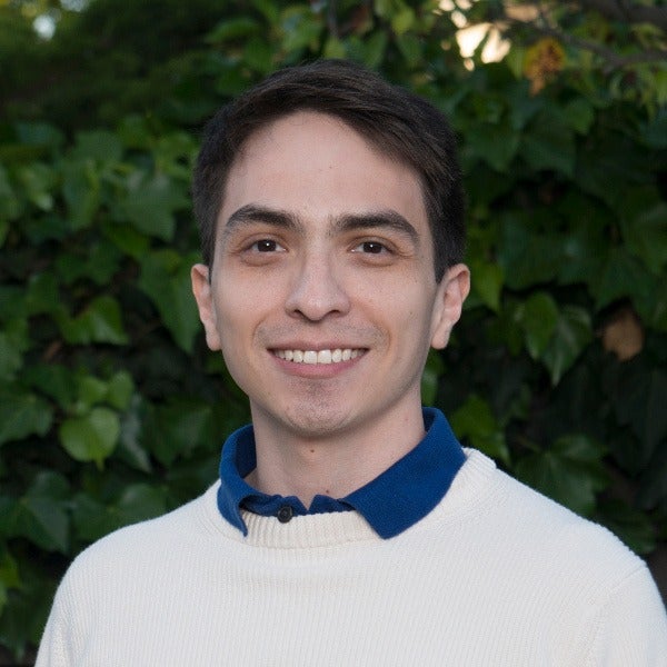 Luis Quiroga, dark brown hair, warm smile, cream sweater over blue collared shirt, green foliage background
