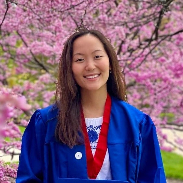 Lydia Kang, long brown hair, warm smile, blue graduation gown with red stole, pink blossoms background