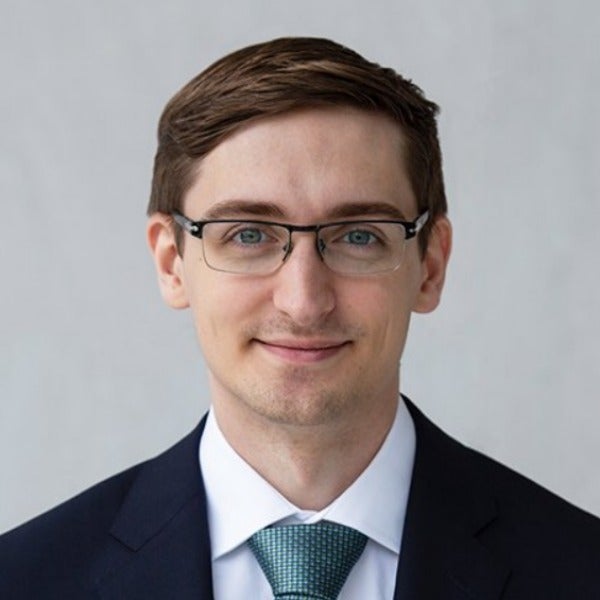 Maclane Speer, brown hair styled short, glasses, light stubble, friendly smile, dark suit with teal tie