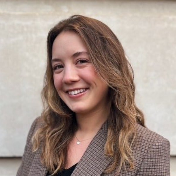 Malea Bauer, shoulder-length brown hair with blonde highlights, warm smile, brown plaid blazer, light background