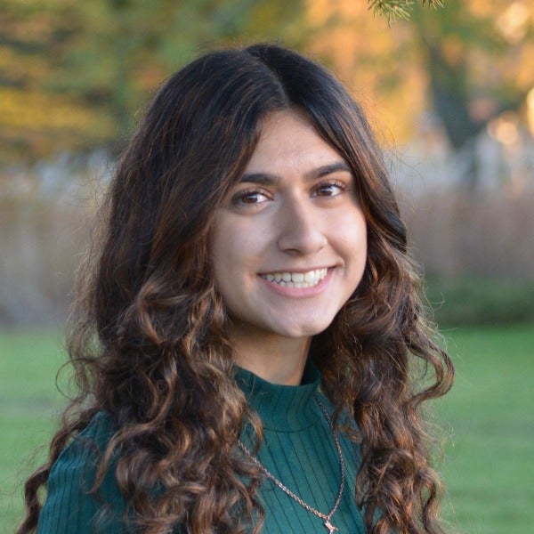 Mandeep Kaleka, long wavy brown hair, warm smile, teal sweater, delicate necklace, outdoor setting