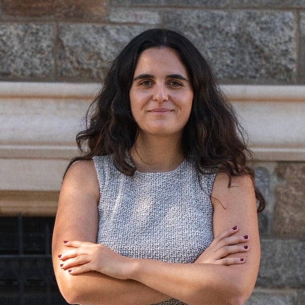 Marta Hernandez, dark shoulder-length wavy hair, arms crossed, gray sleeveless dress, stone wall background