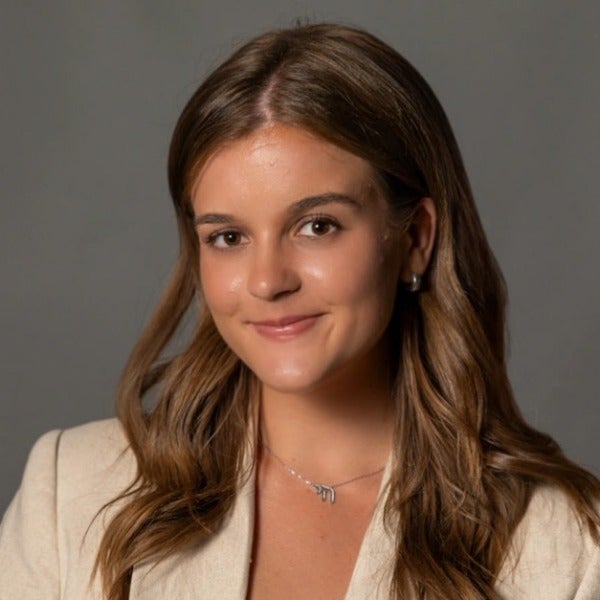 Maya Westerlund, long brown wavy hair, warm smile, beige blazer, delicate necklace, gray background