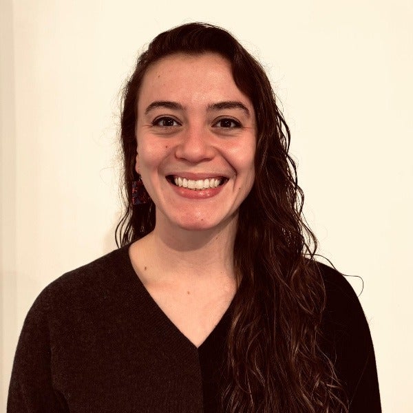 Mia Kazman, long dark brown wavy hair, bright smile, black v-neck sweater, light background