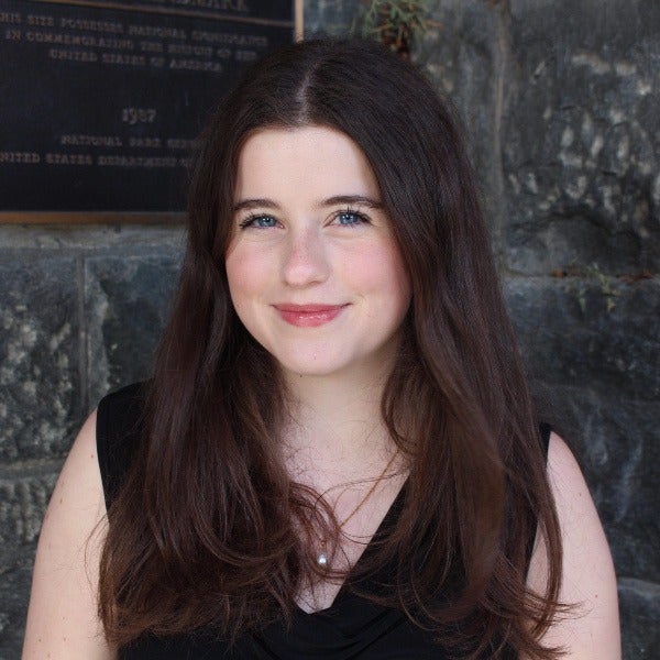 Moira Christ, long brown hair, blue eyes, gentle smile, black sleeveless top, stone wall background