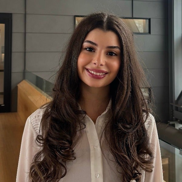 Nashwa Saad, long dark brown wavy hair, warm smile, white button-up shirt, modern office background
