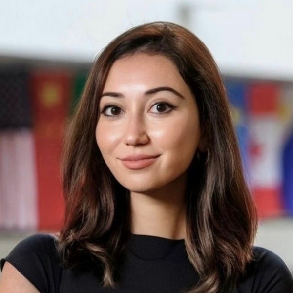 Noora Samadi, long brown hair, warm expression, black top, blurred colorful background