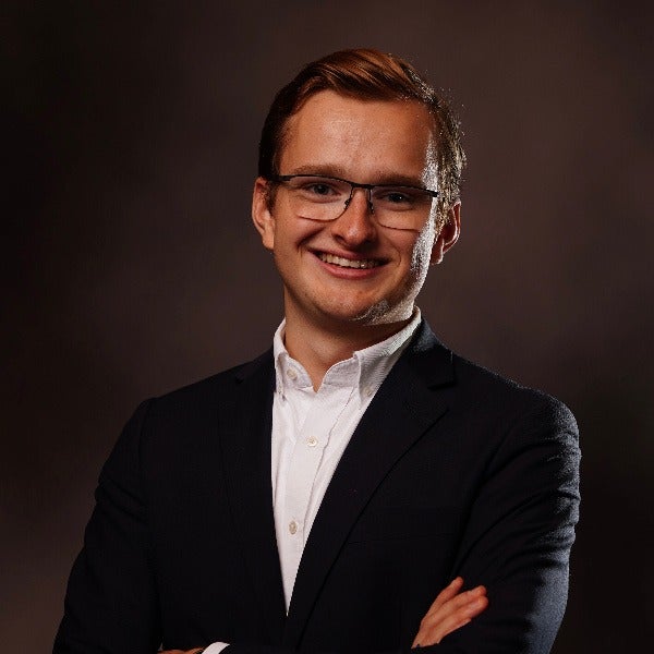Olivier Backx, brown styled hair, glasses, warm smile, black blazer over white shirt, dark background