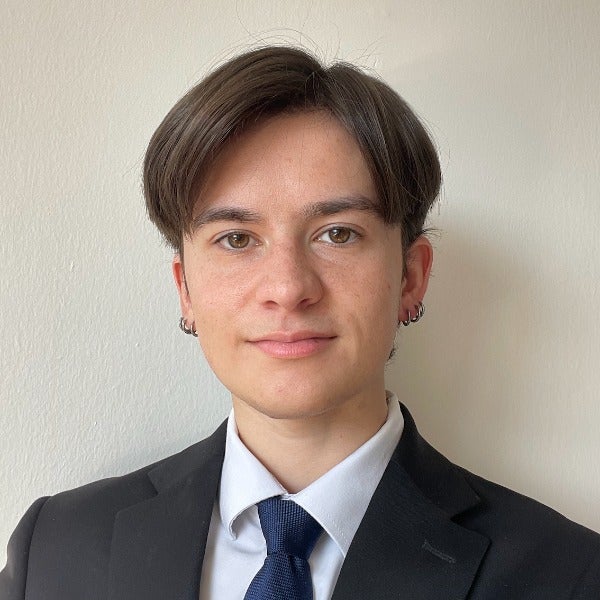 Percy Charlot, brown medium-length hair, earrings, slight smile, black suit with white shirt and blue tie