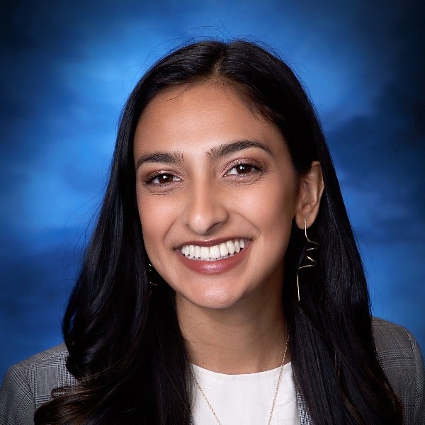 Ritika Saligram, long dark hair, warm smile, gray blazer over white top, blue background