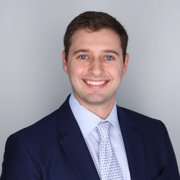 Rylan Forester, short brown hair, blue eyes, bright smile, navy suit, light blue shirt, patterned tie, gray background