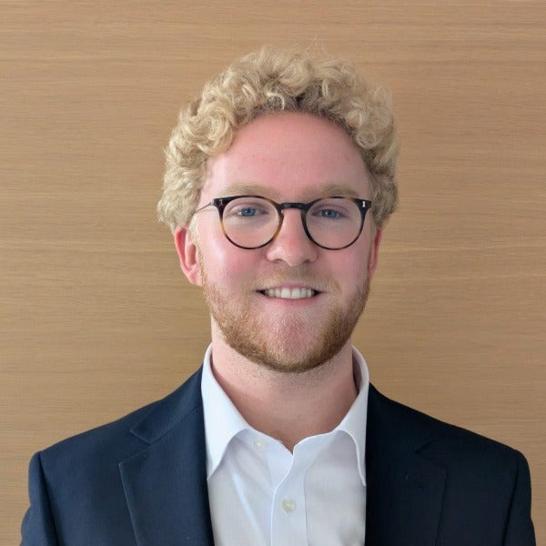 Sandy Robson, blonde curly hair, glasses, light beard, smiling, dark suit jacket, white shirt, wooden background